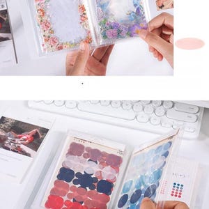 May include: Clear plastic sticker albums with floral and abstract designs. The albums contain stickers in various shapes and colors, including red, blue, and purple. The background includes a white keyboard and other stationery items.