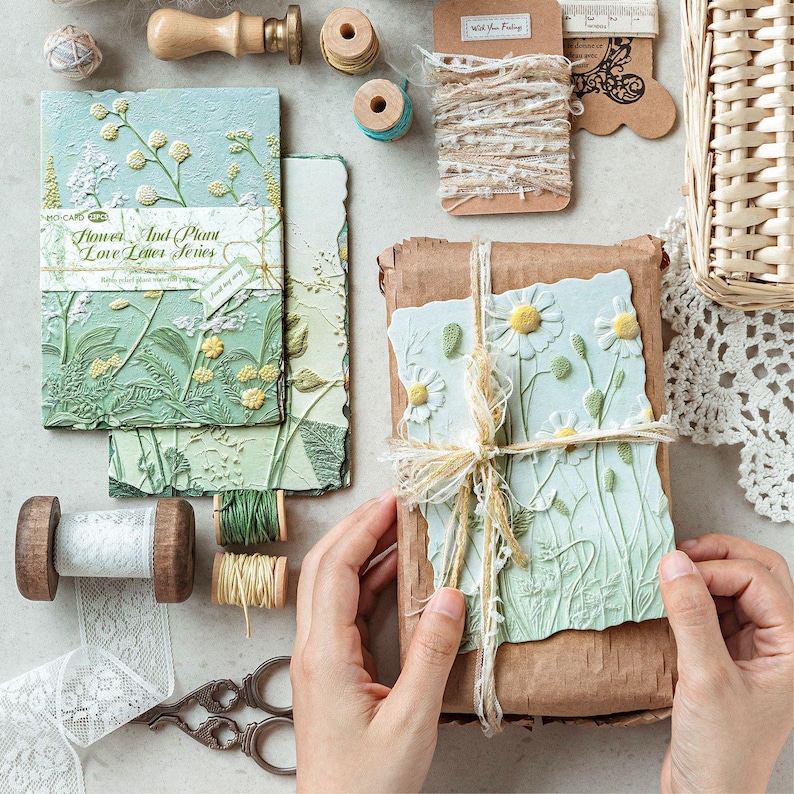May include: A collection of stationery items including textured light green paper cards with embossed floral designs, labelled Flower And Plant Love Letter Series, alongside spools of lace, twine, wooden stamps, and a brown paper-wrapped gift.