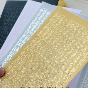 May include: A selection of alphabet stickers in gold, silver, and black. The stickers have small, embossed letters in different fonts. The stickers are arranged in a fan shape, with the gold sticker sheet at the front. The stickers are ideal for crafting.