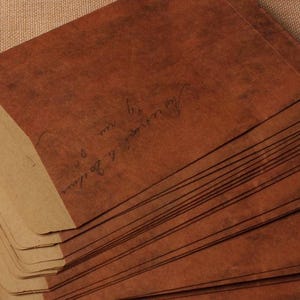 May include: A stack of brown paper envelopes with a lighter tan flap. The envelopes have handwritten script on them. The paper has a textured, aged appearance. The envelopes are stacked on top of each other.