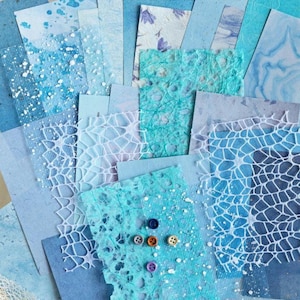 May include: A collection of various blue and white handmade paper sheets. The papers feature different textures, patterns, and shades of blue, including watercolor effects, floral prints, and textured surfaces. Several small, colorful buttons are scattered across one sheet.