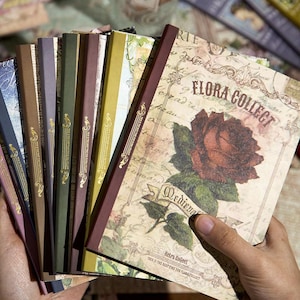 May include: A collection of vintage-style notebooks with various colored spines. The cover of the front notebook features a large red rose illustration with the text "FLORA COLLECT" and "Retro Collect". The notebooks are held in a person's hand.