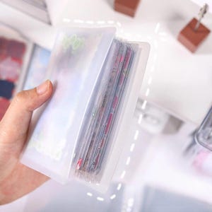 May include: A clear, plastic card holder, partially open, revealing multiple colorful cards inside. The holder is held in a hand, showcasing its design and capacity. The cards appear to be trading cards or similar collectibles.