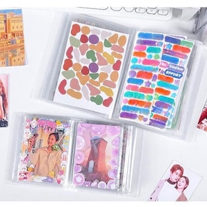 May include: Clear plastic photo album with multiple pages filled with stickers and photos. The stickers are various shapes and colors. The photos feature people and landscapes. The album is on a white surface.