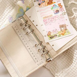 May include: A beige binder with a clear plastic ruler and metal rings. The binder is open, revealing lined paper and colorful illustrations of cartoon characters, teddy bears, and desserts. The ruler is marked in inches.