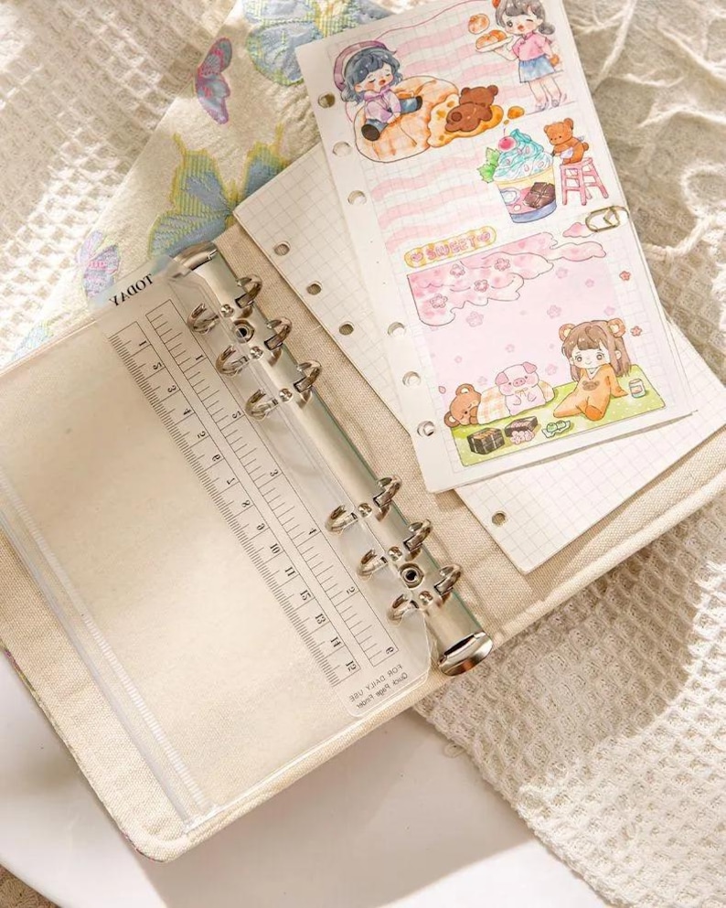 May include: A beige binder with a clear plastic ruler and metal rings. The binder is open, revealing lined paper and colorful illustrations of cartoon characters, teddy bears, and desserts. The ruler is marked in inches.