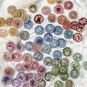 May include: A collection of decorative wax seals in various colors and designs. The seals feature floral, butterfly, and bear motifs. The color palette includes shades of pink, blue, green, and gold. Some seals have the text "E" and "RONTIC".