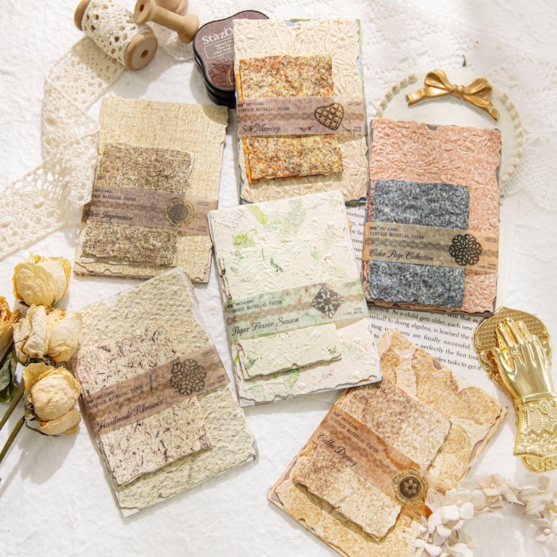 May include: A collection of handmade paper cards with various textures and colors. Each card features a unique design, including floral patterns and decorative accents. The cards are arranged on a white surface, with additional decorative elements.