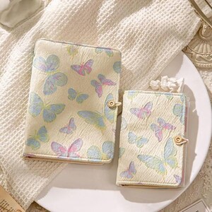 May include: Two cream-colored butterfly-themed notebooks. The notebooks feature a repeating pattern of pastel blue, pink, and purple butterflies. Each notebook has a gold button closure. The notebooks are on a white plate.