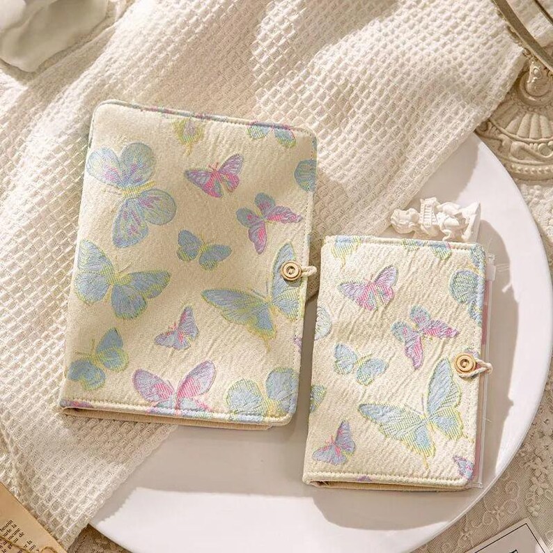 May include: Two cream-colored butterfly-themed notebooks. The notebooks feature a repeating pattern of pastel blue, pink, and purple butterflies. Each notebook has a gold button closure. The notebooks are on a white plate.