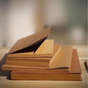 May include: A stack of several brown paper notebooks, with one open to display the interior pages. The notebooks are arranged on a wooden surface, with a soft, blurred background. The paper has a natural, textured appearance.