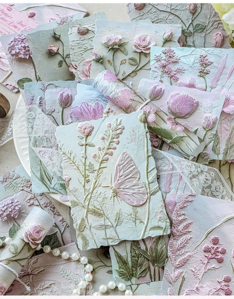 May include: A collection of rectangular paper cards featuring embossed floral and butterfly designs in soft pastel shades of pink, green, and cream. The textured surfaces display raised botanical illustrations, including roses, tulips, and wildflowers, accented by delicate white lace ribbon and a string of white pearls.