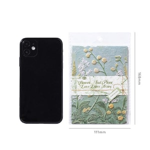 May include: A black smartphone next to a clear plastic package containing stationery cards from the Flower And Plant Love Letter Series. The cards feature embossed floral and botanical designs in shades of green and yellow, measuring 168mm by 111mm.