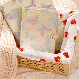 May include: A set of cream-colored notebooks with butterfly patterns in blue and pink, nestled in a woven basket lined with a strawberry-printed fabric. A cream-colored knitted blanket is on the left.