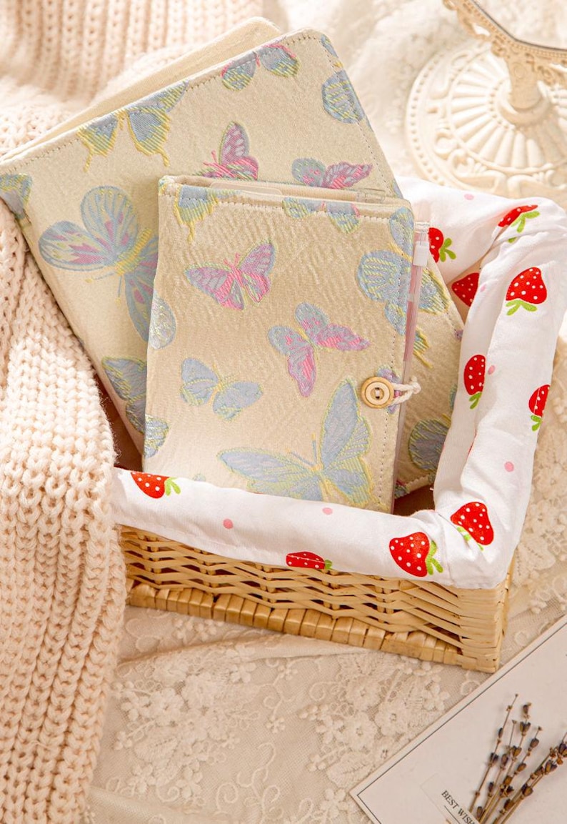 May include: A set of cream-colored notebooks with butterfly patterns in blue and pink, nestled in a woven basket lined with a strawberry-printed fabric. A cream-colored knitted blanket is on the left.