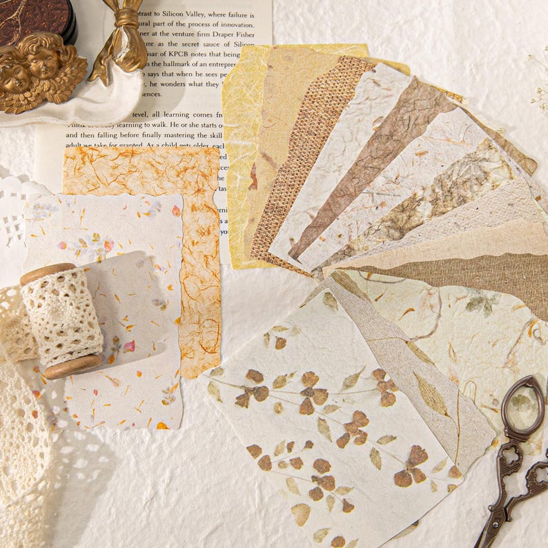May include: A collection of handmade paper sheets in various textures and colors, including beige, brown, and cream. Some sheets feature embedded floral elements. A wooden spool of lace and vintage scissors are also visible, suggesting a craft or stationery theme.