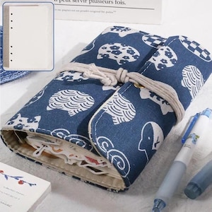 May include: A blue fabric roll-up pen case with a white pattern, tied with a beige cord. The case is open, revealing stickers and stationery. A white notebook and blue pens are also visible.