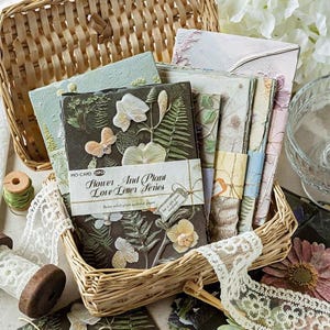 May include: A collection of rectangular paper cards from the Flower And Plant Love Letter Series, featuring embossed botanical and butterfly designs in various colours. The cards are bundled with paper bands and stored in a woven wicker basket.