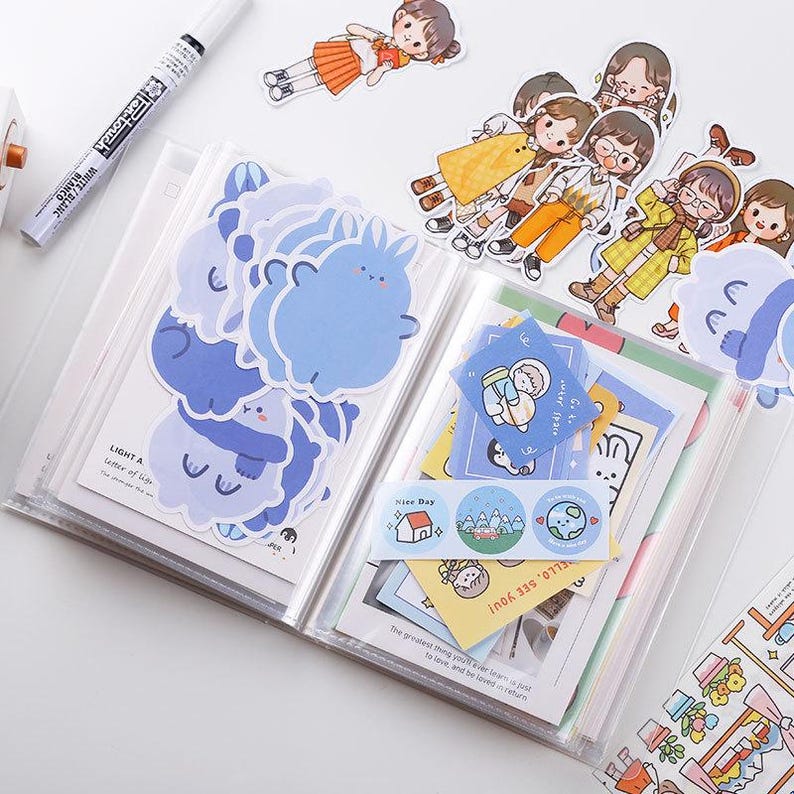 May include: A clear plastic binder filled with stickers and decorative paper. The stickers feature cartoon characters and animals in shades of blue. Additional stickers and illustrations of people are scattered around the binder.