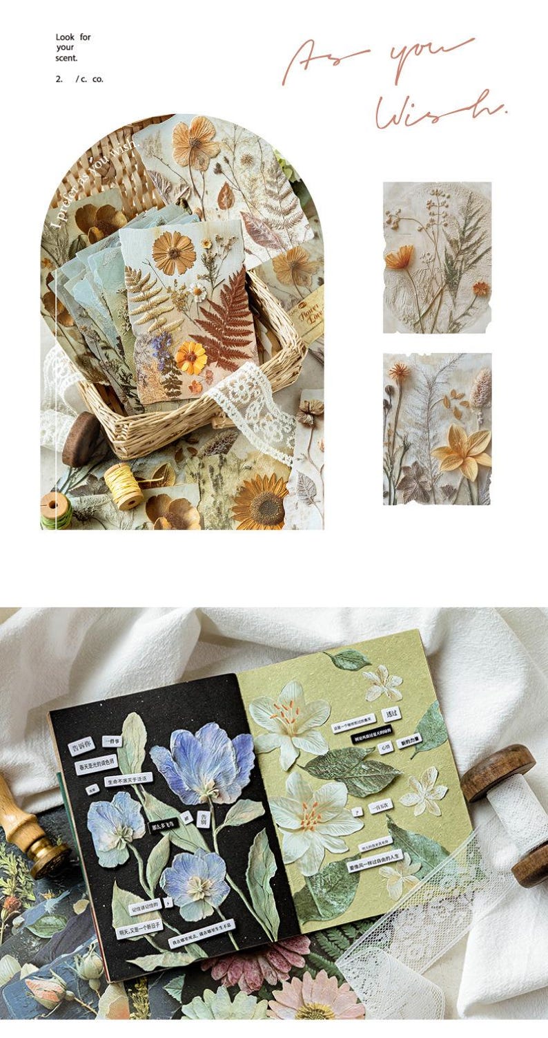 May include: A collection of decorative paper sheets featuring pressed flower and botanical illustrations in warm earth tones and cool blue hues. The set includes rectangular cards with floral arrangements, a notebook with blue and white flower stickers, and text labels.