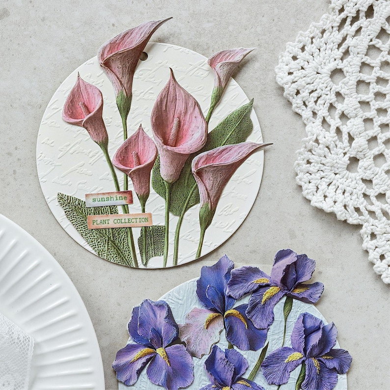May include: Two circular decorative plaques featuring embossed floral designs. The top plaque displays pink calla lilies with green leaves and labels reading sunshine and PLANT COLLECTION. The bottom plaque shows purple irises with yellow centres.