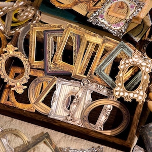 Op de afbeelding: Een verzameling sierlijke fotolijsten in vintage-stijl, in verschillende vormen en maten. De lijsten zijn voornamelijk goud, zilver en bruin, met ingewikkelde details en decoratieve randen. Sommige lijsten zijn ovaal, rechthoekig of cirkelvormig, gerangschikt in een houten bak.