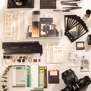 Può includere: Un assortimento di articoli relativi alla fotografia, tra cui rulli di pellicola, strisce di pellicola e un obiettivo della fotocamera. L'immagine presenta scatole di pellicola con il testo "FILM" e "BACKLIGHTING", insieme a vari supporti per pellicola e una fotocamera nera.