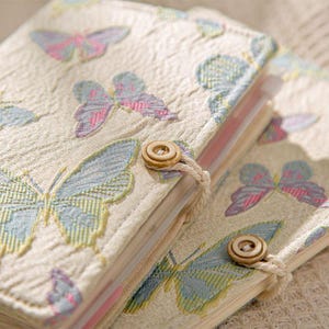 May include: Two beige fabric journals with embroidered butterfly designs in pastel colors. Each journal has a wooden button closure with a matching string. The journals are stacked on top of each other.