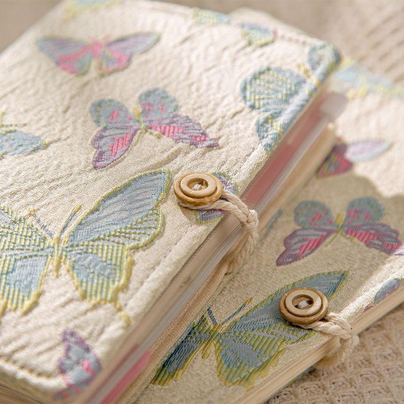 May include: Two beige fabric journals with embroidered butterfly designs in pastel colors. Each journal has a wooden button closure with a matching string. The journals are stacked on top of each other.