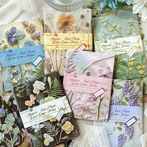 May include: A collection of rectangular paper packs labelled Flower And Plant Love Letter Series, featuring embossed floral and botanical designs in various colours including blue, green, pink, and black, with each pack wrapped in a paper band.