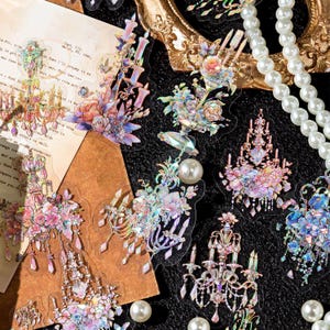 Pode incluir: Uma coleção de autocolantes de candelabro iridescentes com detalhes florais e de pérolas. Os autocolantes apresentam flores rosa, roxas e azuis, com uma estética vintage. Alguns autocolantes são sobrepostos em papel vintage.