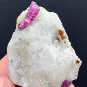 Può includere: Una formazione di cristalli di rubino naturale grezzo. Il rubino è di un vibrante colore rosso-rosato, su uno sfondo di pietra bianca e bianco sporco. Sono visibili anche piccole macchie verdi e marroni.