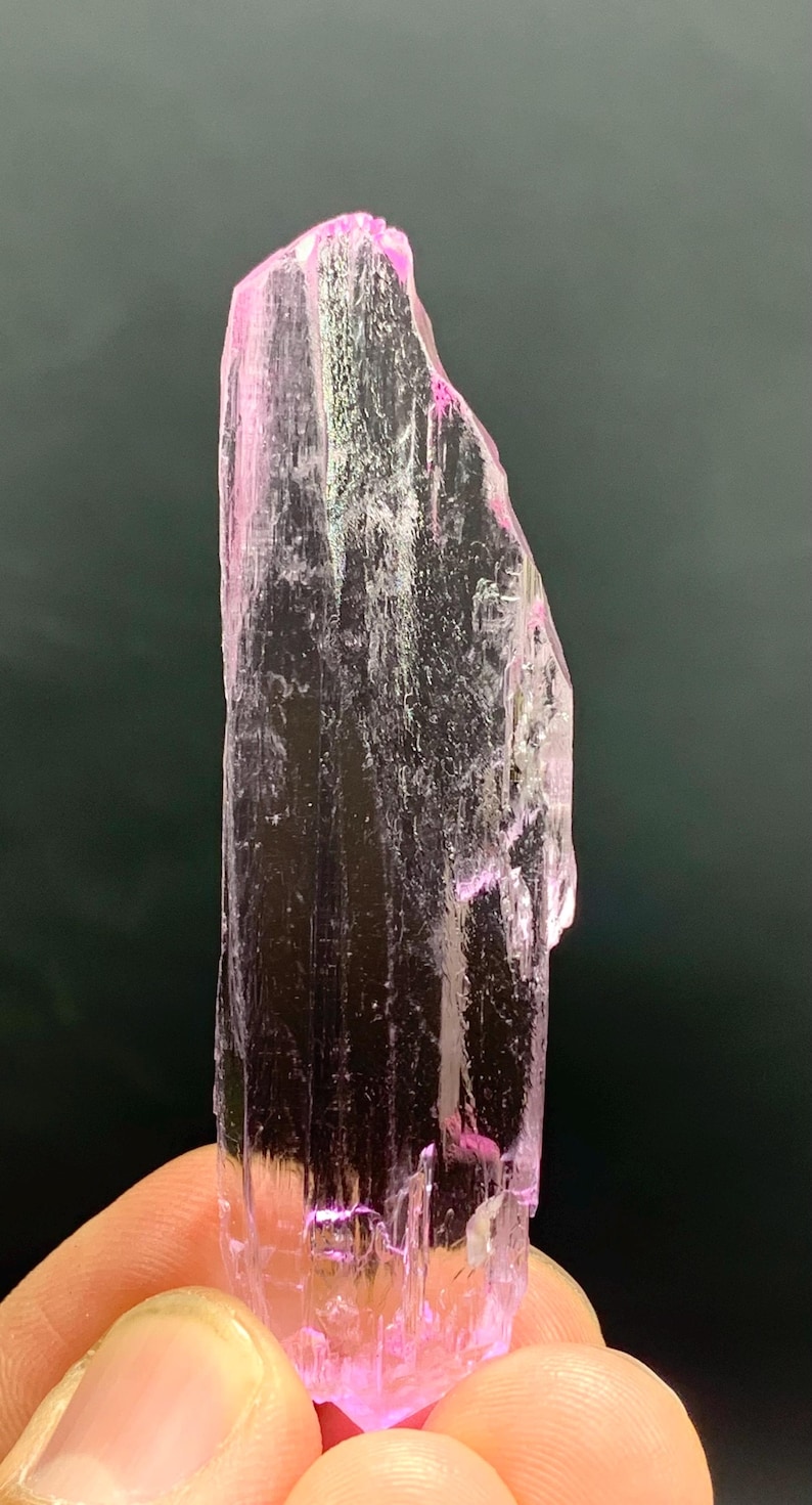 Pu&ograve; includere: Un cristallo rosa traslucido con una punta appuntita e striature su tutta la superficie. Il cristallo &egrave; tenuto in una mano.