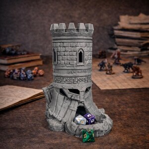 May include: A gray, castle-themed dice tower with a stone-like texture. The tower has a crenelated top, two small windows, and the text "Finn Castr". Several multi-sided dice are visible at the base of the tower, with miniature figurines in the background.