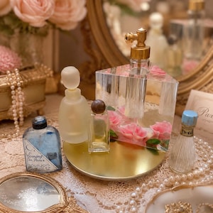 May include: A collection of vintage perfume bottles on a gold circular tray, including a blue glass bottle labeled Evening in Paris Bourjois, a frosted glass bottle, a small clear bottle with a brown cap, and a clear block containing pink roses.