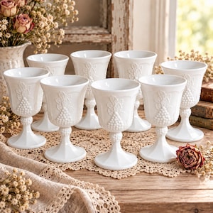 May include: A set of seven white milk glass goblets, each featuring a raised relief pattern of grape clusters and leaves on the bowl and a textured, beaded stem. The goblets have a flared rim and a wide, stable circular base.