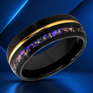 May include: A black band ring featuring a polished gold-tone stripe and an inlay of crushed purple and gold-colored mineral fragments. The ring has a smooth, rounded exterior surface and a dark, reflective finish throughout the metal band.
