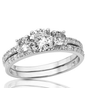 May include: A silver engagement ring set with a three-stone design. The center stone is larger than the two side stones. The band is encrusted with smaller stones. The ring is set on a white background.