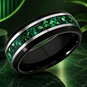 May include: A black ring featuring a central inlay of crushed green stone fragments. The band is bordered by two thin, polished silver-colored metallic stripes that frame the textured green inlay, creating a distinct contrast against the matte black finish.