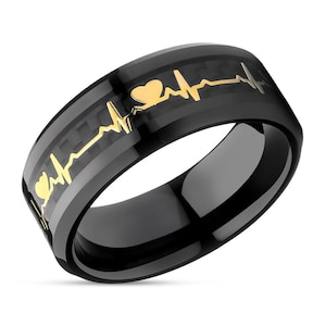 Puede incluir: Anillo negro con un diseño de corazón y latido del corazón en oro. El anillo tiene un patrón de fibra de carbono.