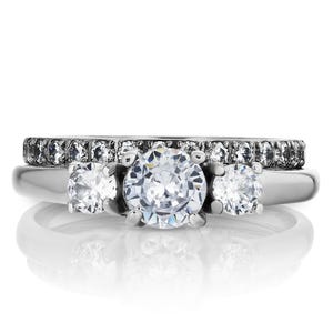 May include: A silver three-stone engagement ring with a larger center stone and two smaller stones on either side. The ring is set with clear round-cut diamonds and has a diamond-encrusted band.