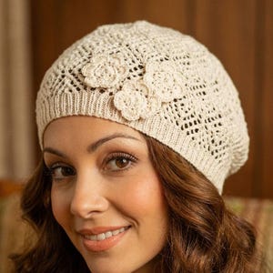May include: A cream-colored, crocheted beret with three decorative flower appliqués. The hat features an open weave design and a ribbed brim. The flowers are a lighter shade of cream, adding a subtle contrast. This stylish beret is a fashionable accessory.