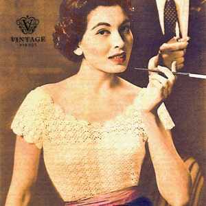 May include: A woman wearing a white crocheted top with a pink ribbon around the waist. She is holding a cigarette holder in her right hand and looking at the camera.