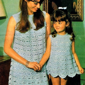 May include: A woman and a young girl wearing matching light blue crochet dresses. The sleeveless dresses have a scalloped hem and a detailed, openwork pattern. The woman is wearing a gold bracelet. The background includes a painting in a gold frame.