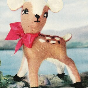 May include: A handmade stuffed deer toy with a brown body, white spots, and a red bow. The deer has white ears, black eyes, and black hooves. The toy is standing on a surface with a lake and mountains in the background.