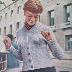May include: A vintage image of a light gray knitted cardigan with four buttons. The cardigan is worn over a patterned top. The image has a retro aesthetic, with a woman smiling in front of a building.