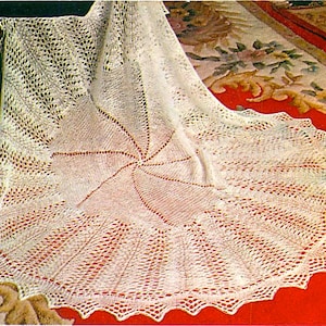 May include: A cream-coloured, finely knitted shawl with a scalloped border. The shawl has a central starburst pattern and openwork details. The delicate fabric is draped, highlighting its flowing form and texture. The shawl is made of a soft material.