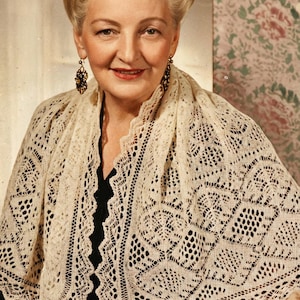 May include: A woman wearing a cream-colored, open-weave shawl with intricate lace patterns. The shawl drapes over the shoulders and features a scalloped edge. The woman is wearing dark earrings and a black top.