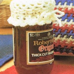 May include: A jar of Robertson's Original Thick Cut Marmalade with a cream-colored crocheted jar cover. The label indicates a net weight of 1 lb. The jar sits on a red mat, with a colorful knitted item in the background.