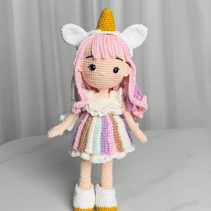 Puede incluir: Una muñeca de unicornio de crochet con pelo rosa, un vestido blanco con rayas arcoíris y zapatos amarillos.